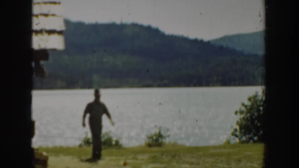 Washington 1940 8mm frame showing a man walking across a grassy lakeshore below wooded hills.