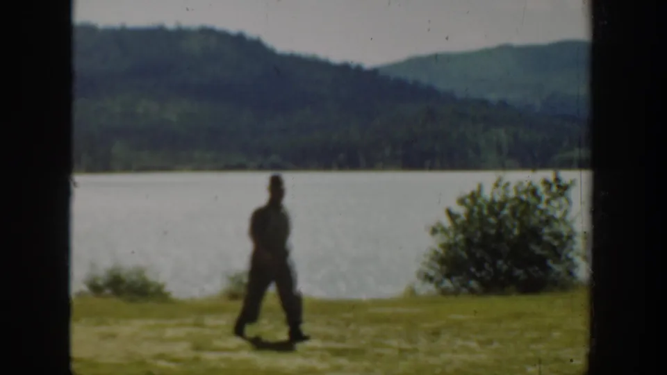 Washington 1940 8mm frame of a man crossing the lakeshore, with water, shrubs, and forested hills behind.