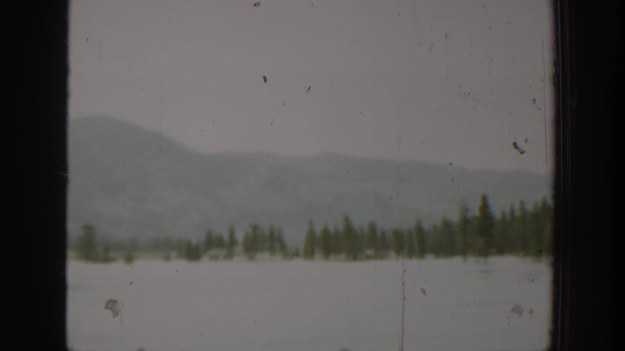 1940 Washington 8mm frame showing pale water, a low evergreen shore, and soft misty hills beyond a dark window edge.