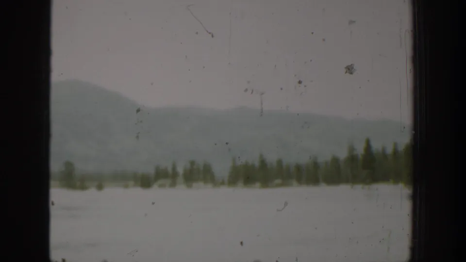 1940 Washington home-movie view of evergreen trees beside pale water, with hazy hills in the distance and window framing at the edge.