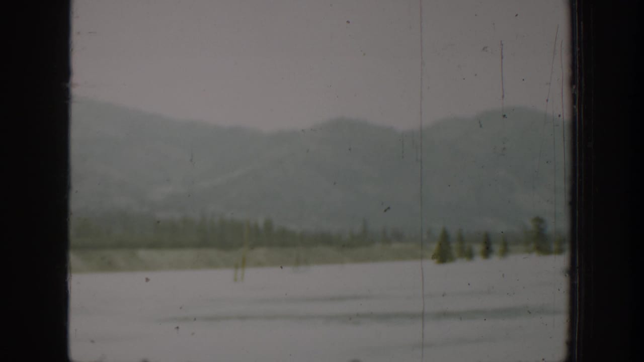 Distant mountains, shoreline trees, and water framed by a car window.