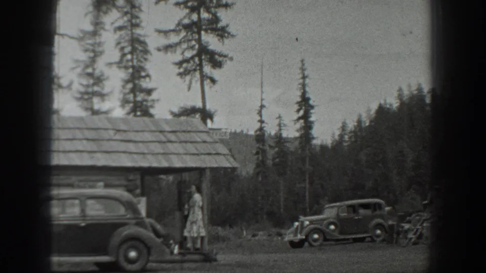 Black-and-white 1940 Washington home-movie frame of a woman near a rustic cabin, with two vintage cars in a wooded setting.