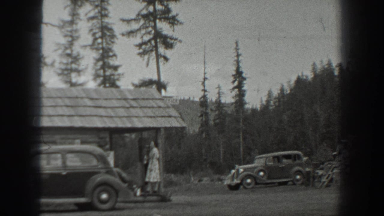 In 1940s Washington home-movie footage, a woman stands near a rustic cabin while two rounded automobiles sit beside tall pines.