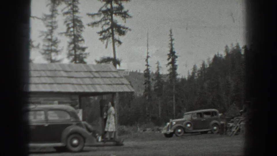 In 1940s Washington home-movie footage, a woman stands near a rustic cabin while two rounded automobiles sit beside tall pines.