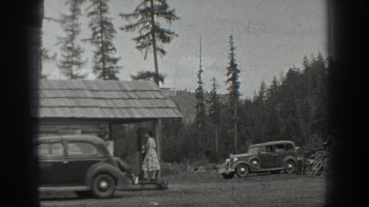 Woman beside a cabin as two vintage cars are parked near a forest clearing.
