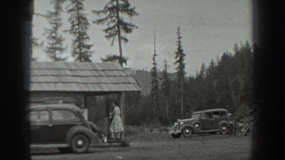 A woman in a light dress stands by a rustic cabin in Washington, with two 1940s-style cars parked in a pine forest clearing.