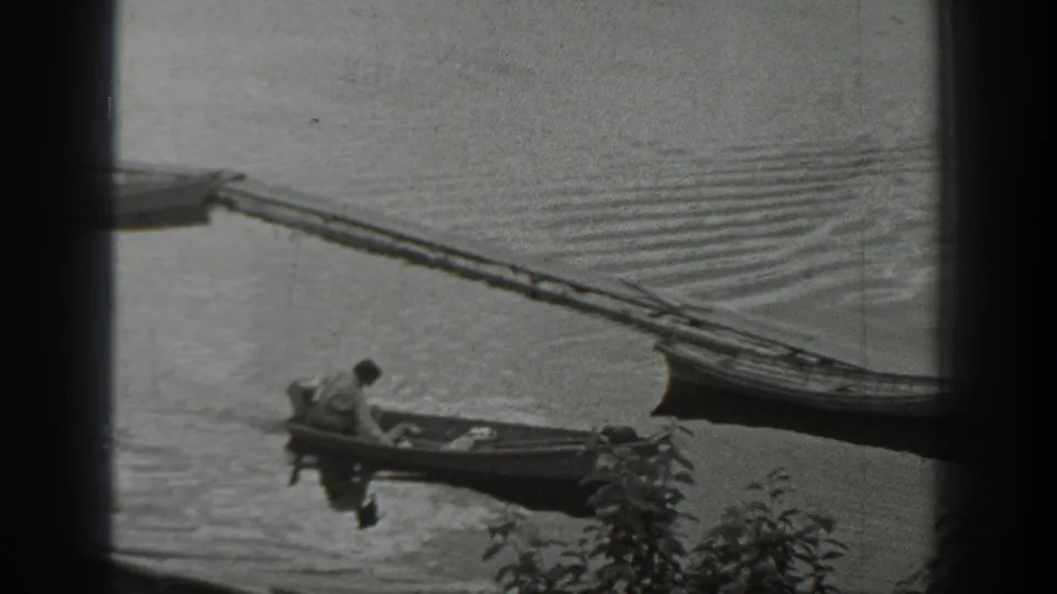 A distant home-movie frame shows the man in a small boat near shoreline plants, with a dock and moored boat behind him.