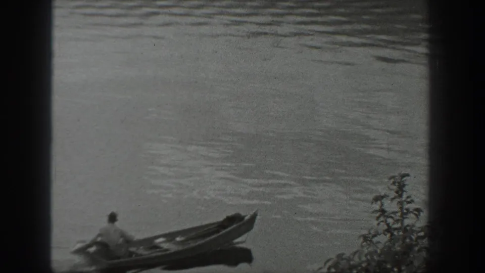 Home-movie view of the canoe passing low in the frame on a calm Washington lake, with a small plant visible at the edge.