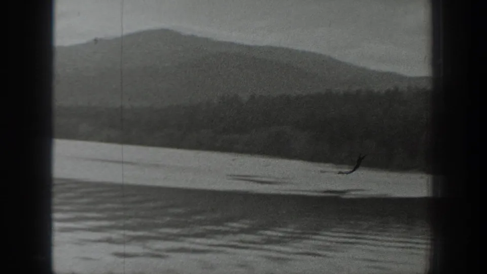 Washington lake landscape in black-and-white 8mm, with rippled water, wooded shoreline, and a low mountain ridge in the distance.