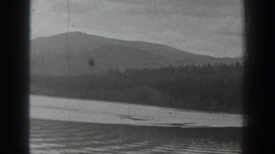 Black-and-white 8mm view of a Washington mountain lake with forested shoreline and hills, partly obscured by dark frame bars.