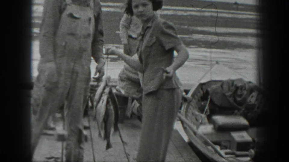 1940s Washington home-movie frame with a woman holding a fish on a wooden lakeside pier while a man in overalls stands nearby.