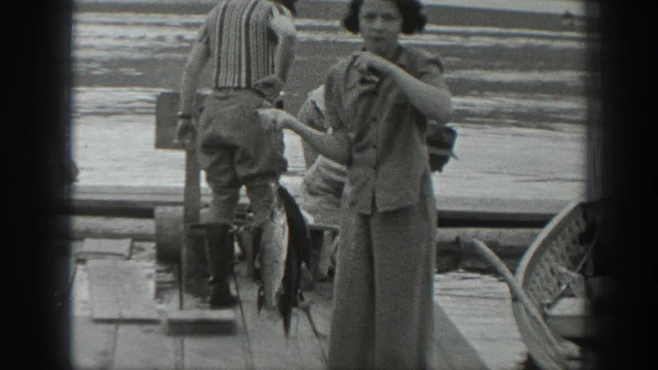 In 1940 Washington home-movie footage, a woman stands on a dock with a large fish beside a striped-shirted man near lake water.