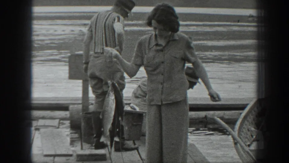 A woman in button-up shirt and trousers stands on a Washington dock near a hanging fish and fishing gear in black-and-white 8mm footage.