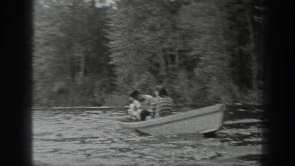 A small open boat carrying seated passengers moves across a forested Washington lake in grainy black-and-white home-movie footage.