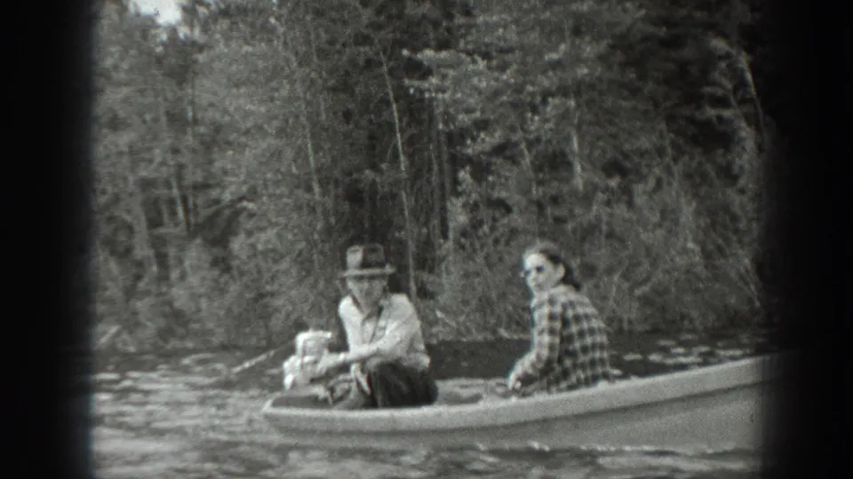 A closer 8mm view shows the woman in plaid and a hat-wearing man seated in the boat near the lake shore.