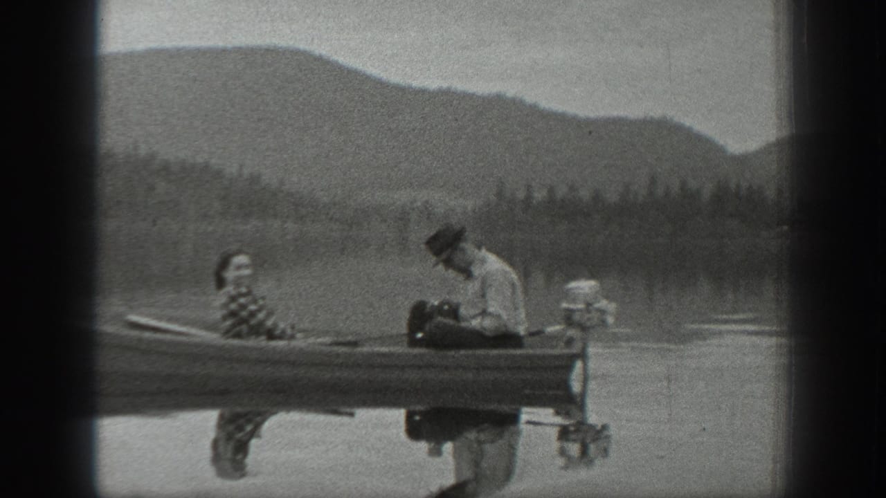 Washington 1940 home-movie frame showing two adults in a small motorboat on a lake with forested hills and mountains beyond.