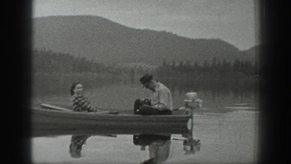Washington 1940 home-movie frame showing two adults in a small motorboat on a lake with forested hills and mountains beyond.