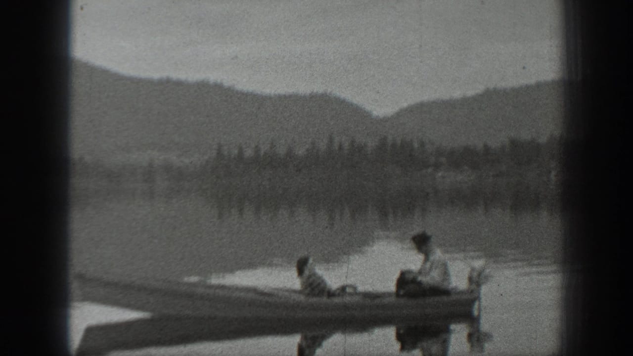 Small boat with two people sits on a calm lake below wooded hills and mountains.