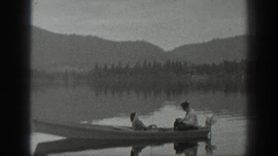 Wide 1940 Washington lake view with two people in a small boat, tree-lined shore, and mountains in the distance.