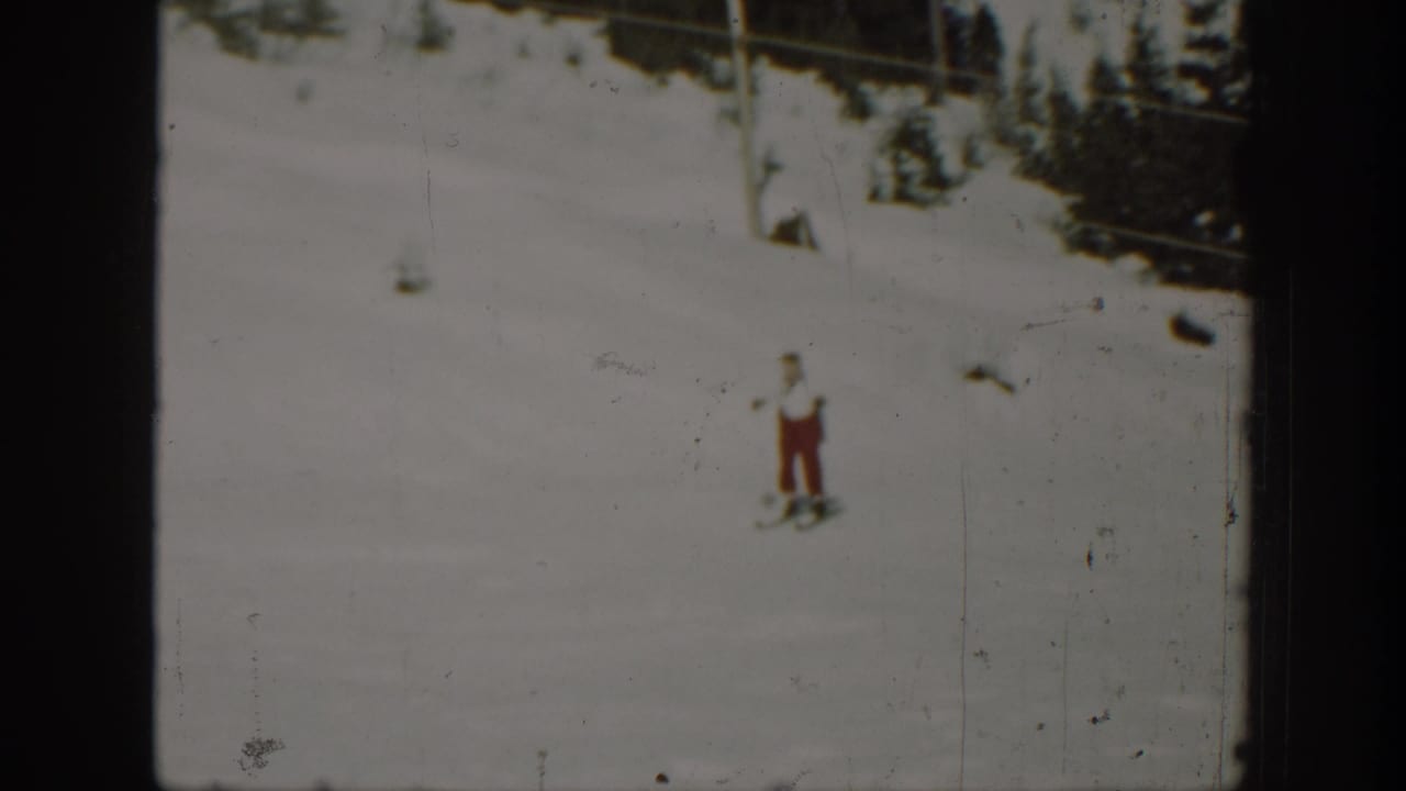 1940 Washington 8mm frame showing a distant adult skier in red pants on a snowy hillside below dark trees.