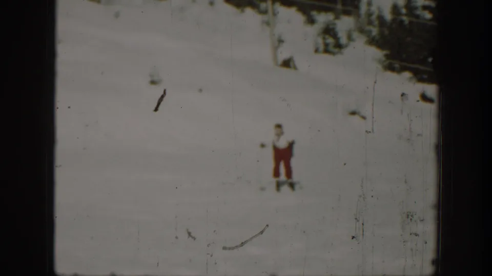 1940 Washington home-movie frame with a small adult skier crossing a pale snowfield near a dark wooded edge.