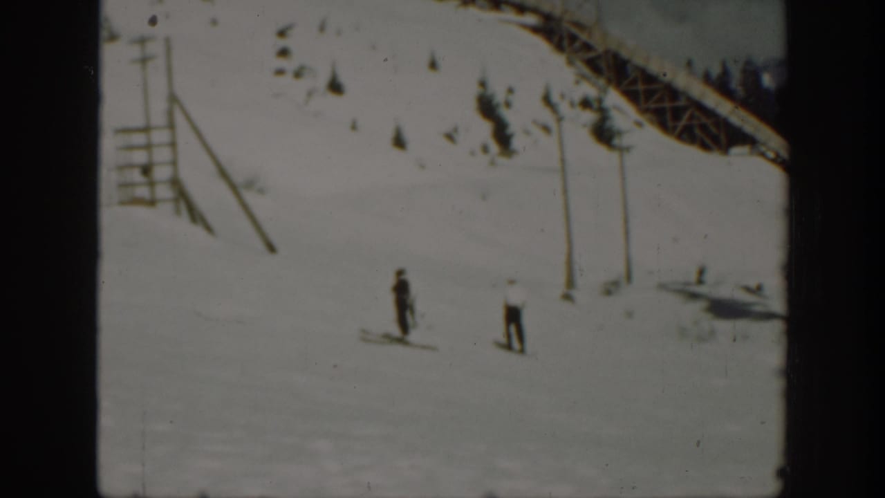 1940 Washington home-movie frame showing two distant skiers on a bright snow run with a lift-like structure at the upper edge.