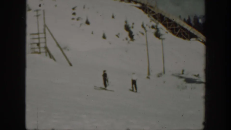 1940 Washington home-movie frame showing two distant skiers on a bright snow run with a lift-like structure at the upper edge.