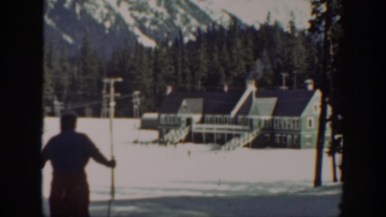 1940 Washington 8mm frame showing a skier with poles crossing a snowy resort area toward a lodge and forested mountains.