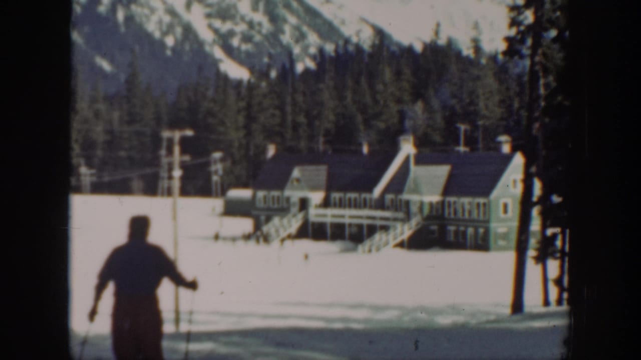Skier glides farther from the camera toward the lodge across bright snow.