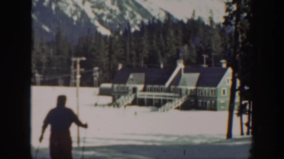 1940 Washington resort footage frame with the skier moving away, framed by tree trunks, lift poles, lodge roofs, and mountains.