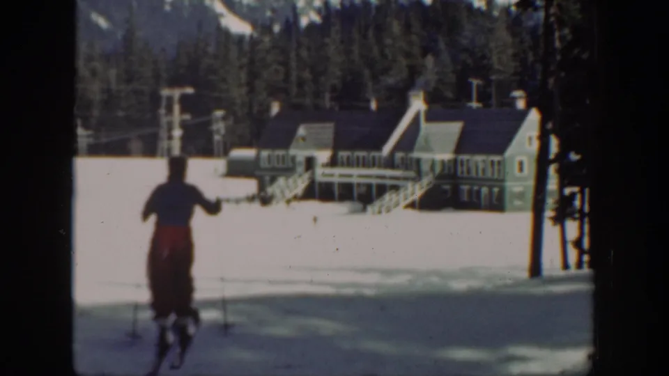Washington 1940 home-movie frame of a skier from behind, with resort lodge steps, lift infrastructure, evergreens, and mountains visible.