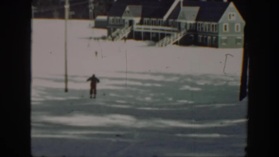 The skier continues across the snow at a distance, with wooden houses, stairs, and a utility pole behind.