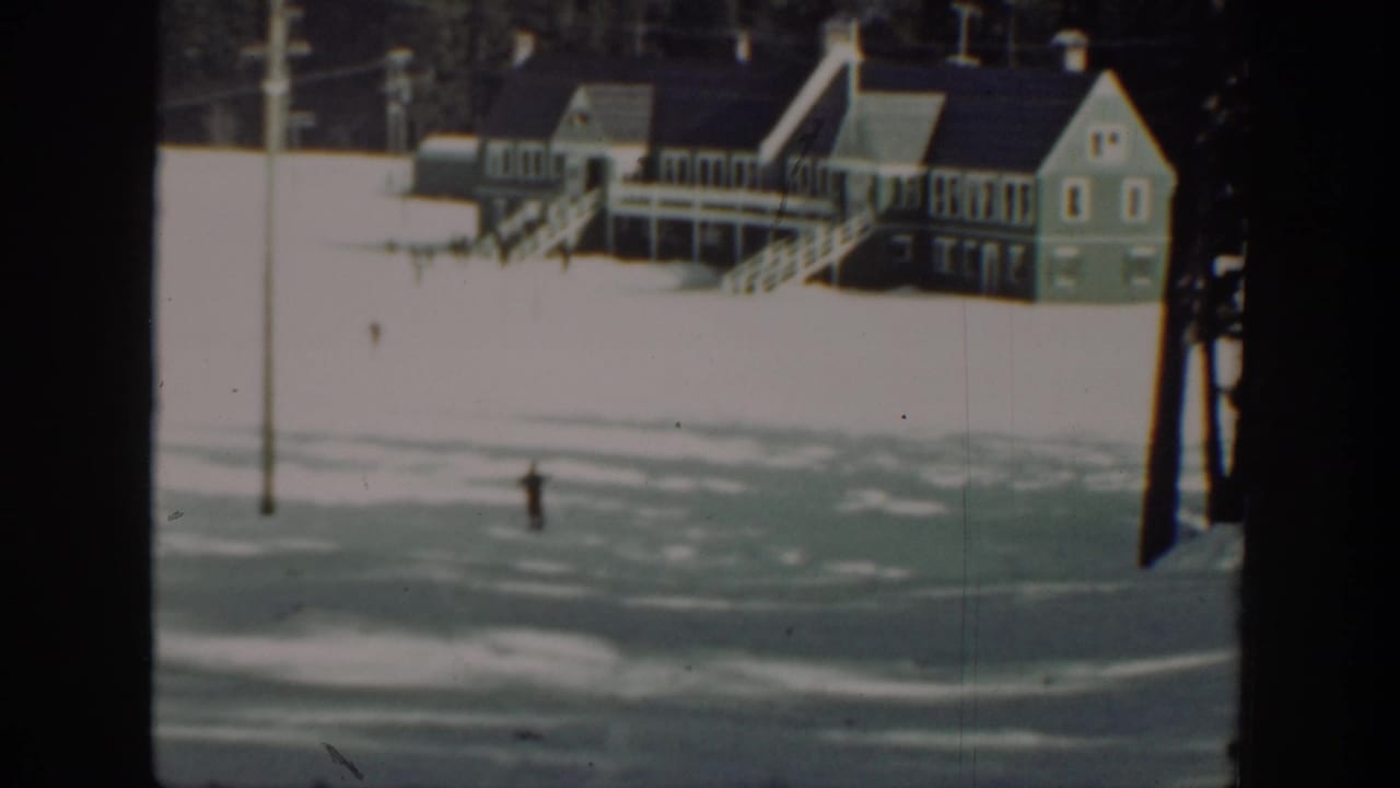 Small skier moving across snow with a lodge-like building in the background.