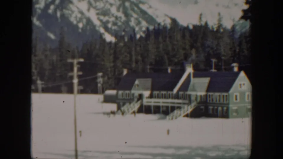 A 1940s Washington winter frame shows the lodge-like building, snowy foreground, evergreens, and mountains beyond.