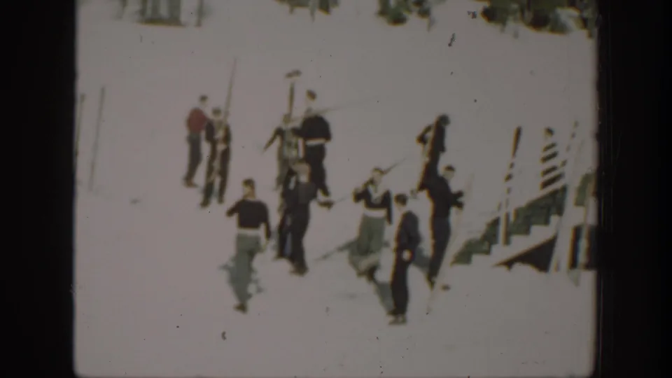 A home-movie view of people moving through a snowy ski area, with skis, poles, and a striped resort-side structure partly visible.