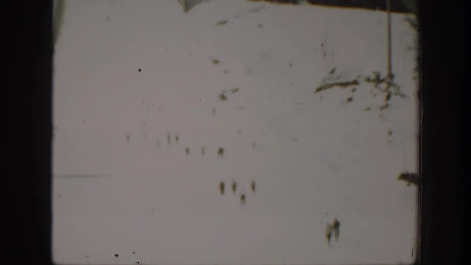Bright overexposed snow scene with small distant figures and scattered ski or track marks in the frame.