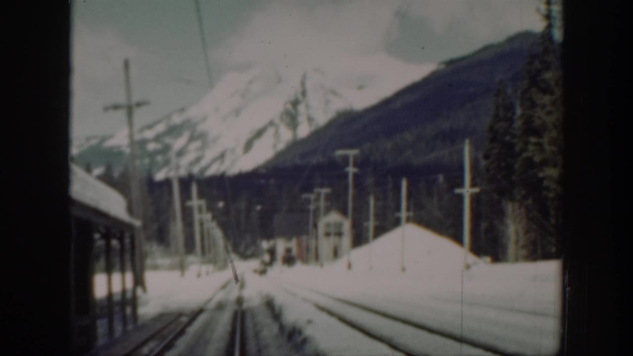 Snowy railway tracks beside a train with utility poles and mountains beyond.