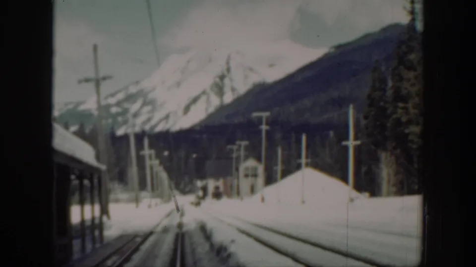 Washington 8mm view from a moving train showing snowy tracks, utility poles, forested slopes, and snow-capped mountains.