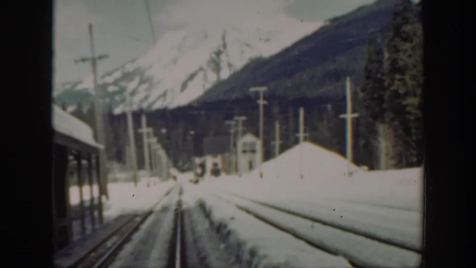 Washington mountain railway scene with snow-covered tracks, a small station structure, utility poles, and a train edge at left.