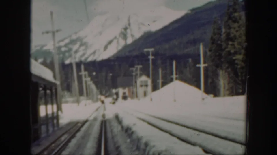 Soft 8mm Washington frame of a snowy railway corridor with poles, a small station building, forested slopes, and mountains.