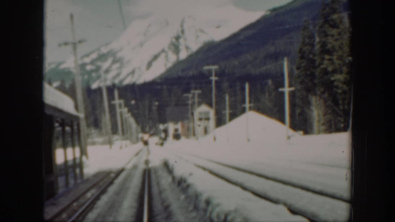1940s Washington rail footage looking forward along snow-covered tracks toward mountains, with a train car edge at left and utility poles beside the line.