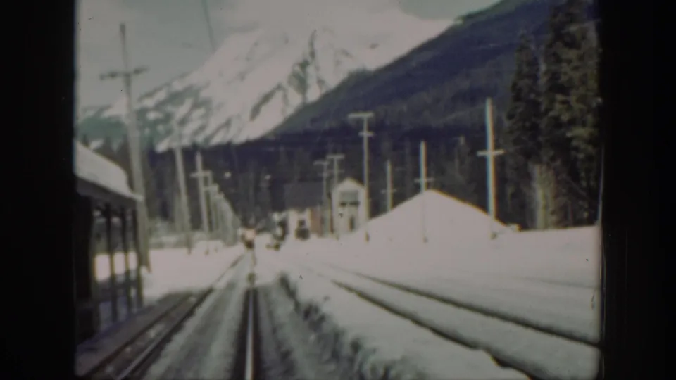 1940s Washington rail footage looking forward along snow-covered tracks toward mountains, with a train car edge at left and utility poles beside the line.