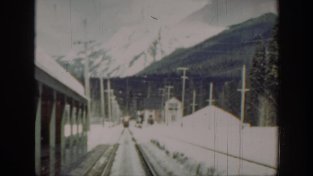 Snow-covered tracks run toward a small structure with mountains and poles in the background.