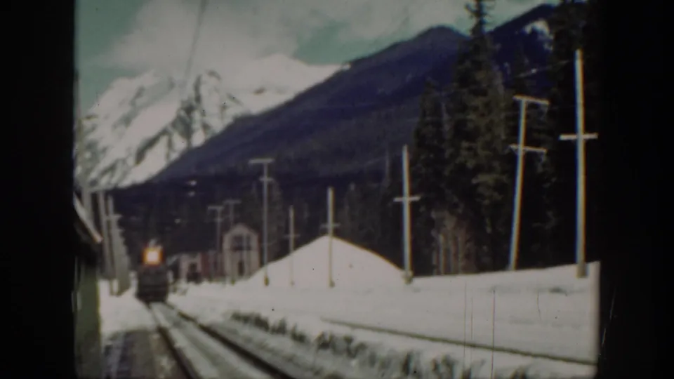 1940s Washington home-movie frame showing an oncoming locomotive with a glowing headlight on snow-lined tracks beneath mountain slopes.