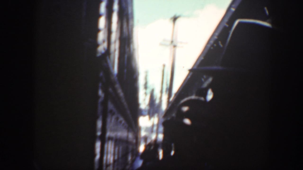 1940 Washington home-movie frame looking through a narrow rail-side passage, with dark silhouettes, utility poles, and blown-out daylight.