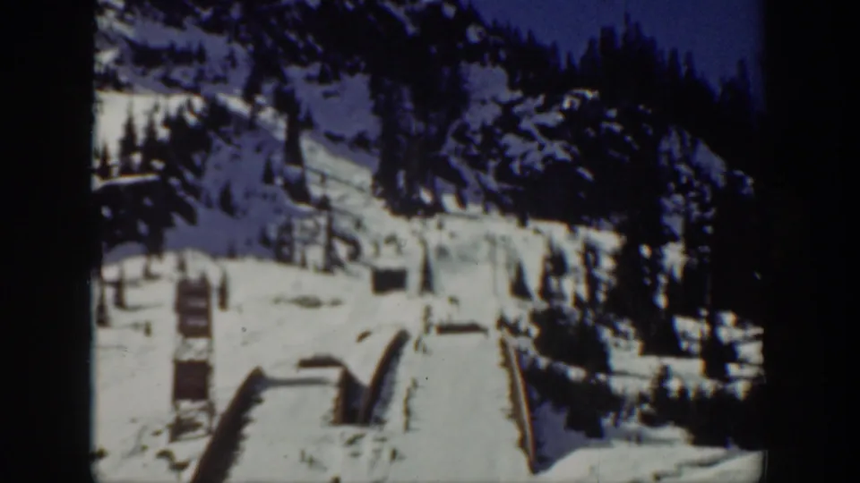 1940 Washington 8mm frame showing a wide winter mountain scene with pine-covered slopes, snow structures, and distant people.
