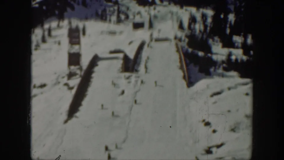 1940 Washington frame of a snowy ski slope with simple wooden ramp features and dark evergreen trees above.