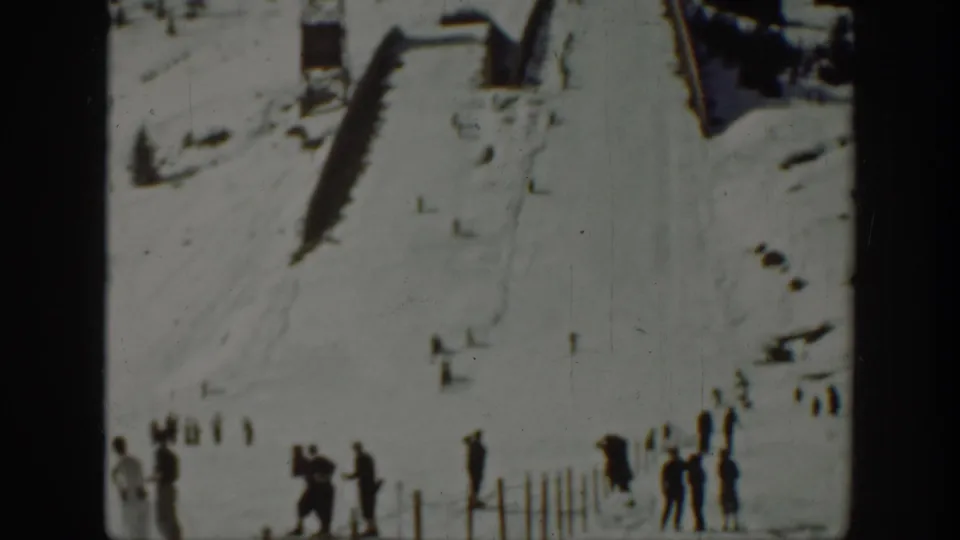 1940 Washington home-movie frame with distant spectators by fence posts below a snowy slope and wooden ramp structures.