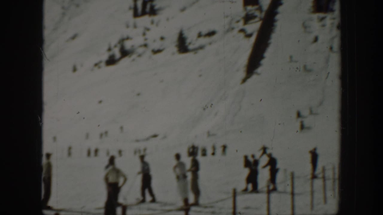 Several people stand on snow near a fence while skiers move in the background.