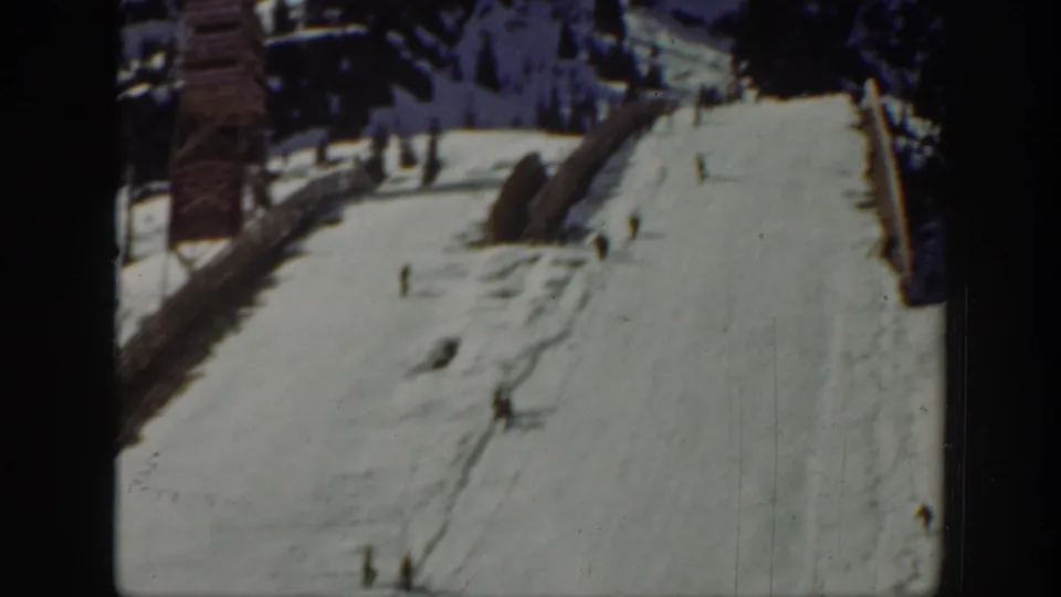 1940 Washington ski-area frame showing a broad snowy run with distant skiers, tracks, and side structures.